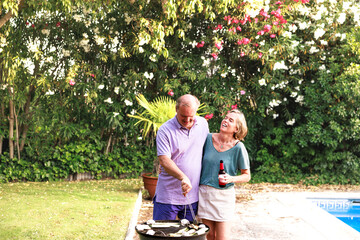 Happy senior couple grilling vegetables in garden barbecue next to the pool