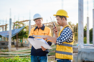 An elderly man and a middle-aged Asian man, both wearing hard hats and reflective vests, closely...
