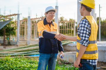 An elderly engineer and a middle-aged man shake hands at a construction site, symbolizing mutual...