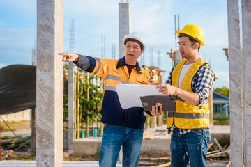 An elderly man and a middle-aged Asian man, both wearing hard hats and reflective vests, closely...