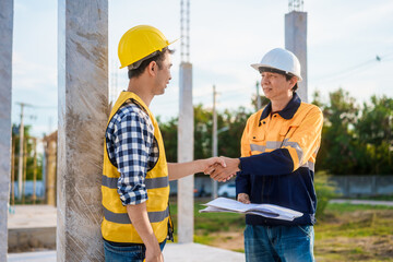 An elderly man and a middle-aged Asian man, both wearing hard hats and reflective vests, closely...