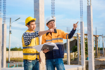 An elderly man and a middle-aged Asian man, both wearing hard hats and reflective vests, closely...