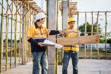 An elderly man and a middle-aged Asian man, both wearing hard hats and reflective vests, closely...