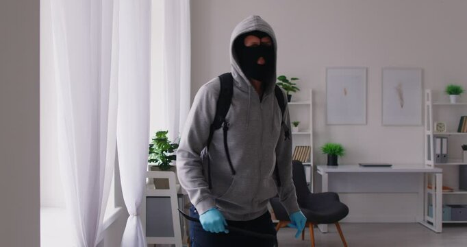 Burglar breaks into a house or an apartment. Robber in a balaclava, a hoodie and gloves, with a crowbar and a backpack enters home through a window, looks around and walks inside the room