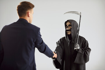 Contract with death. Serious creepy Grim Reaper and young man in suit shake hands after making...