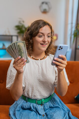 Successful rich woman holding smartphone and waving dollar bill money fan sitting on sofa couch at...