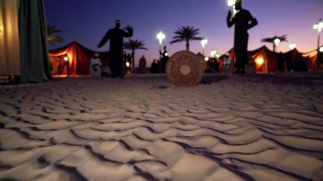 Large traditional bedouin drum rolling on desert sand with blurred silhouettes of musicians and illuminated tents in the background at dusk