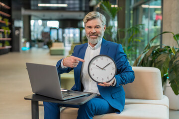 It is your time. Mature boss businessman with laptop showing time on wall clock, approve, pointing...