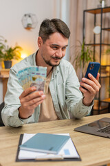 Planning family budget. Smiling man counting money Ukrainian hryvnias UAH cash use smartphone...