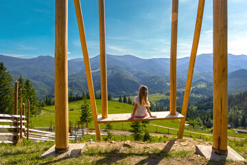 Young girl on swing enjoying mountain view. Child sits on a large wooden swing, looking out at...
