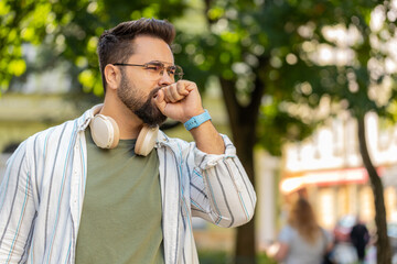 Caucasian young adult man tourist coughing while standing on city street. Sick male suffers from...