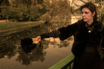 A confident and sophisticated lady in a strict black suit of the 19th century posts with a fan against the backdrop of an park.