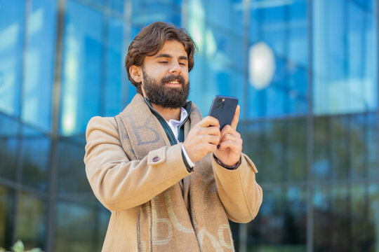 Indian Arabian man tourist chatting on internet on mobile phone standing outdoors. Happy smiling young Lebanese guy using cellphone typing email message communicate by SMS on downtown city street.