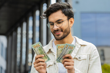 Happy joyful Indian man counting money cash holding dollars banknotes bills budget count salary profit reward bribe outdoors. Successful rich Arabian Hindu guy in downtown city street. Business people