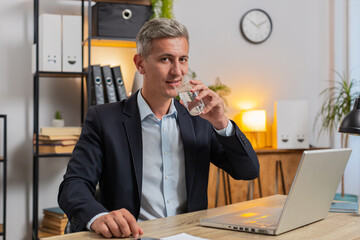 Focused Caucasian businessman sitting at workplace desk drinking water while working with laptop in...