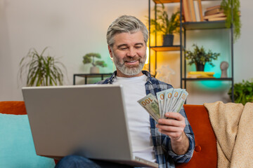 Happy Caucasian senior old man sitting on sofa counting money cash and using laptop calculate...