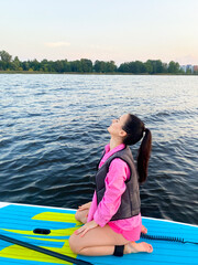 Peaceful Young Woman in Pink Outfit Meditating on Paddleboard SUP at Sunset on Calm Lake. Vertical