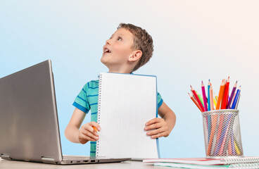 A cheerful young boy shows his notebook during an online learning session, sitting at a desk with a laptop and colorful pencils, ready for a fun and creative school day