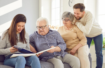 Old, senior parents and adult children happily look through a family photo album together at home....