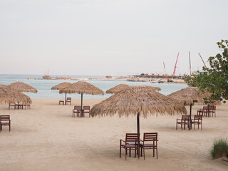 A serene Doha beach with straw umbrellas, traditional boats, and a skyline of modern buildings.