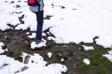 A hiker steps on a patch of snow on a muddy stream in a winter landscape, showing the contrast between the melting snow and the green grass below, hiking in the mountains in winter