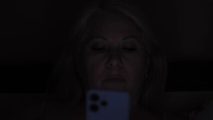 portrait of a beautiful caucasian woman 45 years old in the dark lying in the bedroom using a smartphone chatting or using social networks, woman with a phone in bed at night - Powered by Adobe
