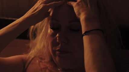 portrait of middle aged caucasian blonde woman giving herself a head massage with her hands while lying in bed in the evening, massage against headache and migraine