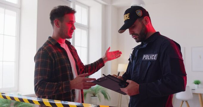 Police man officer talking to witnesses making interview standing with young man robbery victim after burglary interrogating him at fenced crime scene. Robbery, safety of property concept. 4k video.