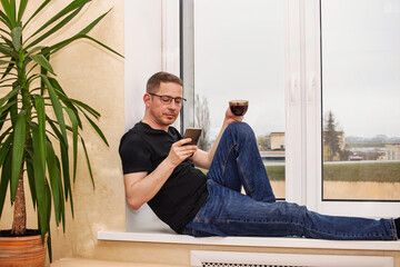 Relaxing man enjoys coffee while using smartphone by the window on a cloudy day