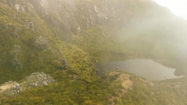 Steep volcanic walls draped in green drop toward a tranquil pool at the heart of Mount Apo as morning fog gleams gold during a slow aerial sweep.
