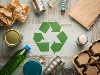 Sorting recyclables with cans, glass, and paper products arranged around a green recycle symbol on a wooden table