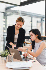 Two young women working together on laptop in modern office, focused and engaged in discussion with documents and stationery on table, bright natural light