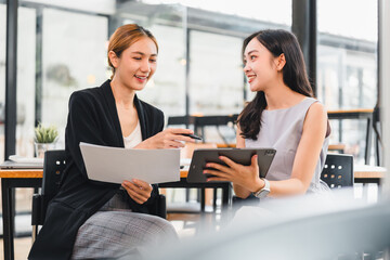 Two young women discussing business document and digital tablet in modern office, smiling and collaborating with positive energy