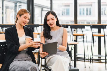 Two young women sitting in modern cafe, discussing work on digital tablet with coffee cup, showing collaboration and focus in bright, casual setting