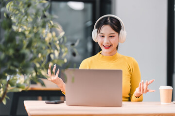 Young woman wearing headphones using laptop at table with coffee cup and smartphone, enjoying music...