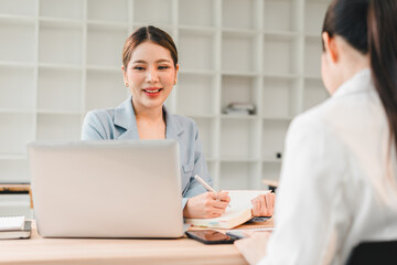Obraz premium Two businesswomen having meeting in modern office, one woman smiling and taking notes while using laptop, professional discussion with positive atmosphere