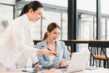 Two businesswomen working together in modern office, discussing project with laptop and notebook,...