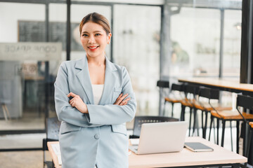 Confident young businesswoman standing with arms crossed in modern office, smiling warmly with...