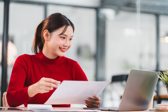 Young woman in red sweater working with documents and laptop in modern office, smiling and focused on task, bright and productive atmosphere