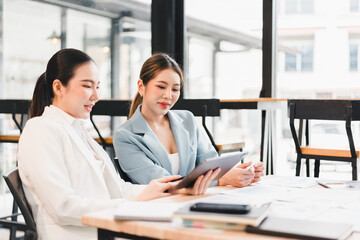 Two businesswomen discussing work while sitting at table with documents and tablet in modern office, showing focus and collaboration in bright environment