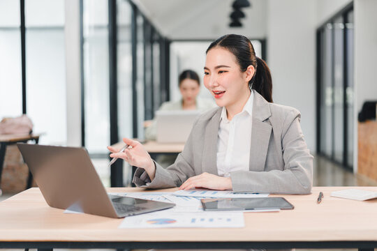 Young businesswoman working on laptop in modern office, smiling and engaged in video call, with documents and tablet on desk, bright workspace with glass walls and natural light