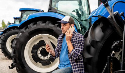 Modern farmer making phone call and using digital tablet near farm tractors at equipment dealership