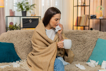 Young woman at home wrapped warmly in plaid blanket holding hot tea sitting on sofa feeling sick....