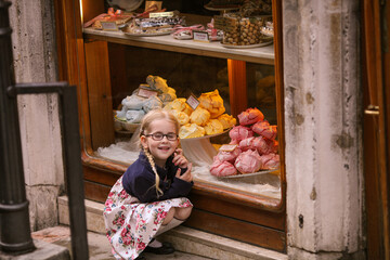 Dreamy blond girl with glasses crouches near candy shop, sweetshop, confectionery, pastry shop in...