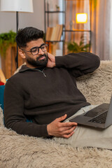 Indian man at home massages sore neck while sitting in front of laptop looking tired after long...