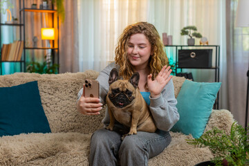 Young Caucasian blogger woman waves hand during video call conversation on home sofa, smiling and...