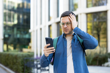 A concerned man outside is looking at his smartphone, hand on his head, possibly in response to bad...
