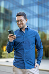 A man in glasses is using a smartphone outside in front of a modern office building, he seems pleased.