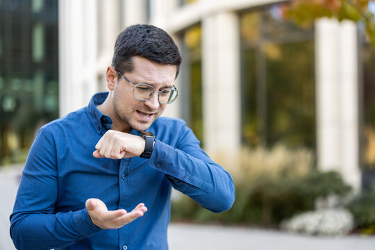A man wearing glasses uses a smartwatch to talk using an app on the street in front of a building