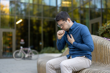 A man coughs and holds his chest while seated outdoors. Blurred background shows a building, bench,...